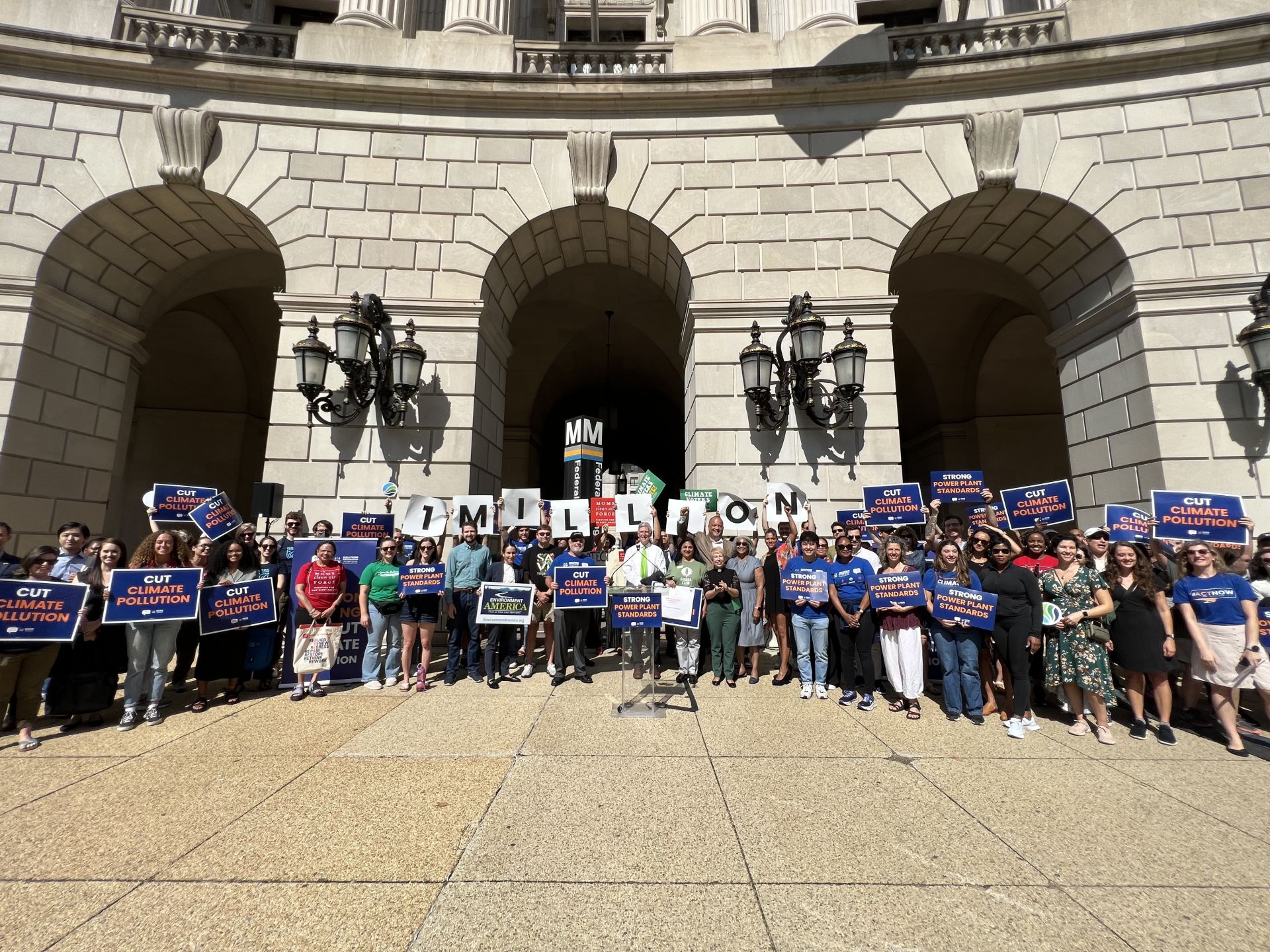 Image of a few dozen people standing in a crowd outside a stone building with arches, holing signs that read "1 MILLION"